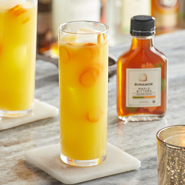 A glass of orange liquid with ice next to a bottle of Runamok Orange Maple Bitters.