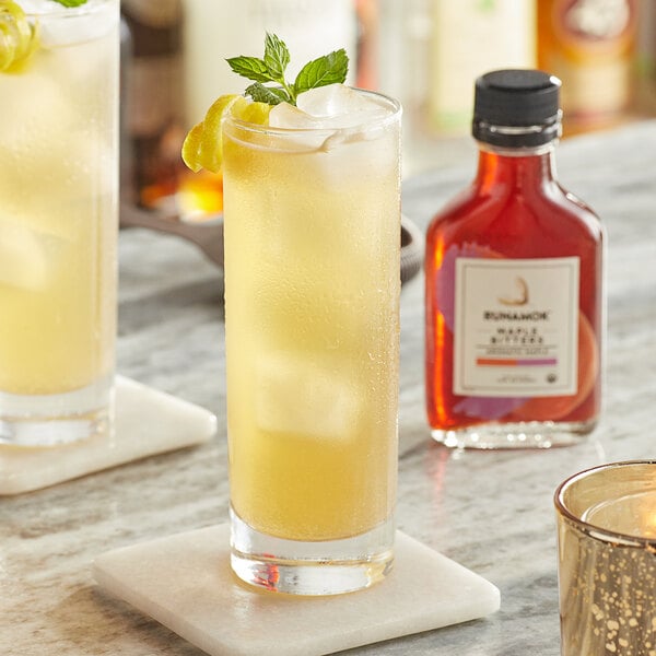 A glass of yellow liquid with ice and a mint leaf with a bottle of Runamok Aromatic Maple Bitters.