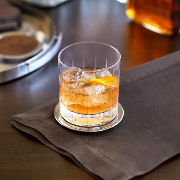 A clear glass rocks or double old fashioned glass with a patterned design, filled with an iced beverage and garnished with an orange peel, placed on a napkin.