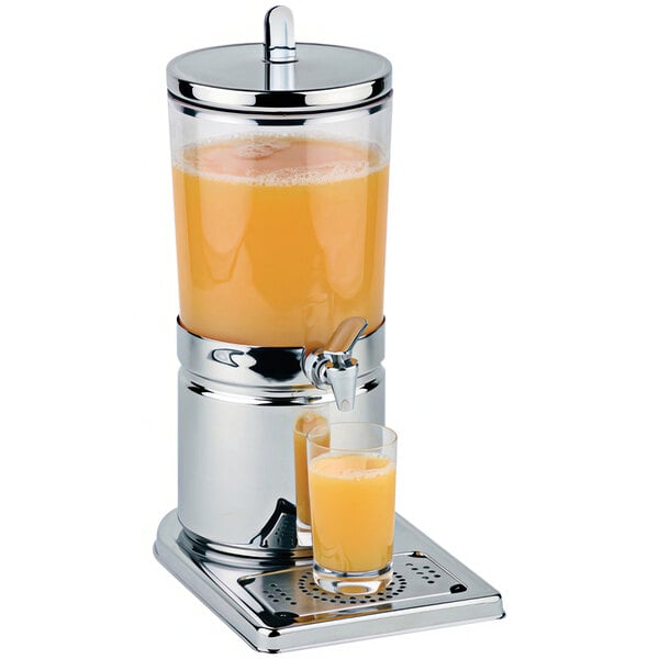 A stainless steel APS beverage dispenser filled with orange juice on a stand next to a glass of orange juice.