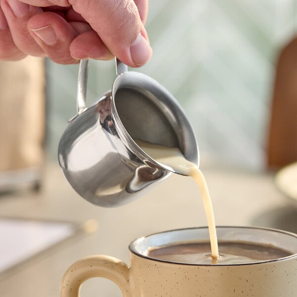 A stainless steel bell creamer pouring cream into a mug of coffee.