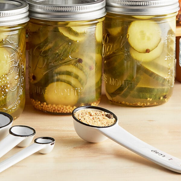 A Ball jar of pickles next to a white measuring spoon filled with pickle mix.