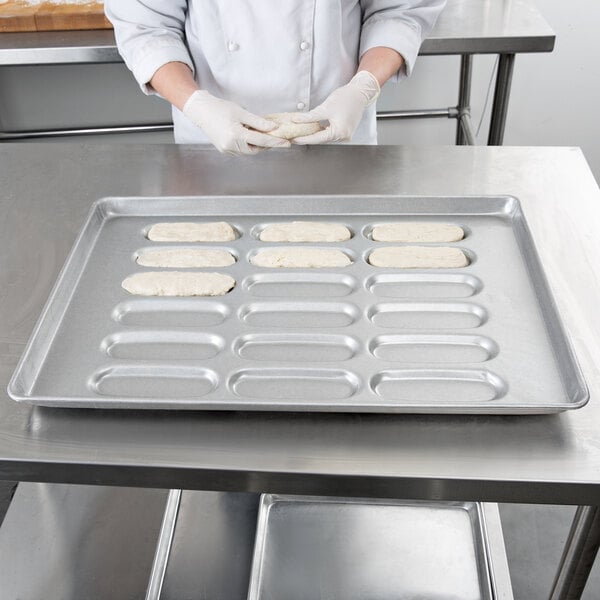 A person in a white uniform holding a Chicago Metallic hot dog bun pan filled with dough