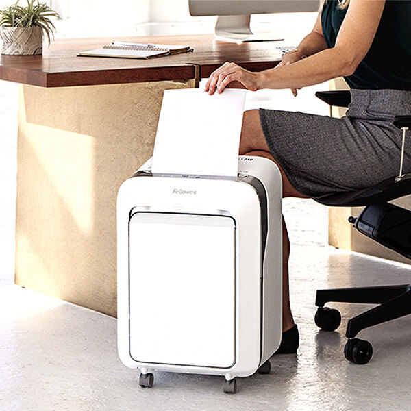 A woman sitting in a chair using a Fellowes white micro-cut shredder to shred a piece of paper.