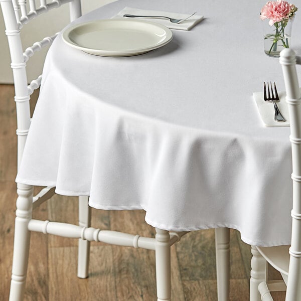 A white table with a Choice white round table cover and a white plate with a fork on it, with flowers in the background.