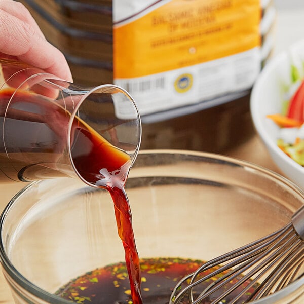 A hand pouring brown Colavita Balsamic Vinegar into a bowl of liquid.