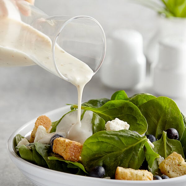 A bowl of fresh spinach salad with croutons, blueberries, and cheese being topped with creamy Italian dressing from a glass container.