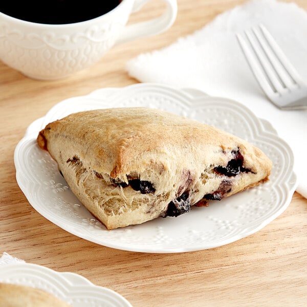 A plate with a David's Cookies blueberry scone on it with a white cup of coffee.