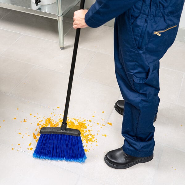 A warehouse broom with blue flagged bristles and a 48-inch handle being used to sweep debris on a tiled floor.