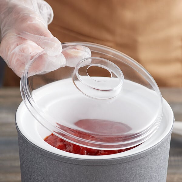 A gloved hand using a Carlisle clear plastic lid with a hole to cover a container of ketchup.