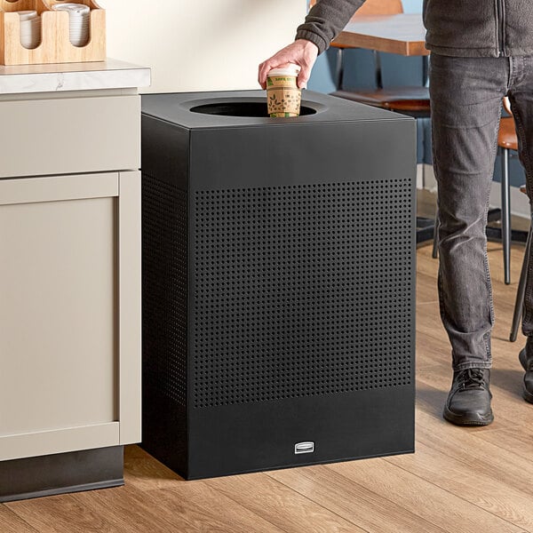 A man standing in front of a Rubbermaid black steel designer square trash can on a table in a coffee shop.