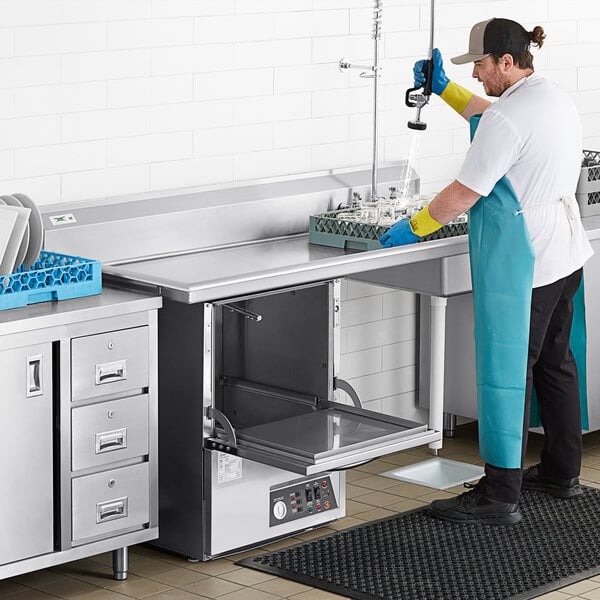 A man in a blue apron washing dishes on a stainless steel Regency dishtable in a professional kitchen.