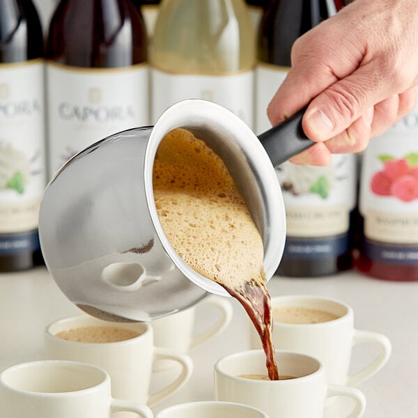 A person pouring coffee into a white cup from a stainless steel Turkish coffee decanter.