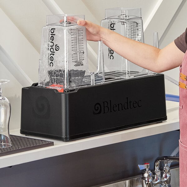 A Blendtec rapid rinser station on a counter with a hand holding a measuring device over a glass of water.