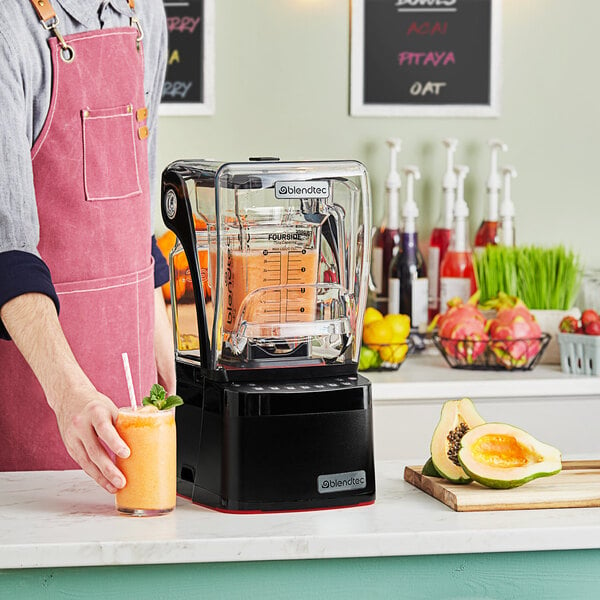 A man in an apron holding a glass of orange juice next to a Blendtec commercial blender.