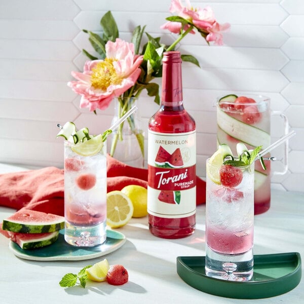 A close up of a Torani Puremade Watermelon Flavoring Syrup bottle next to a glass of watermelon vodka and lemonade.