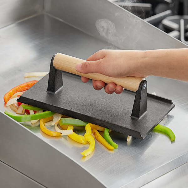 A hand using a Choice cast iron steak weight with vegetables in a pan.