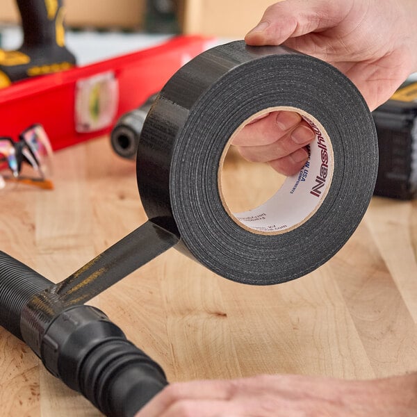 A roll of black Nashua duct tape being applied to a pipe fitting on a wooden workbench.