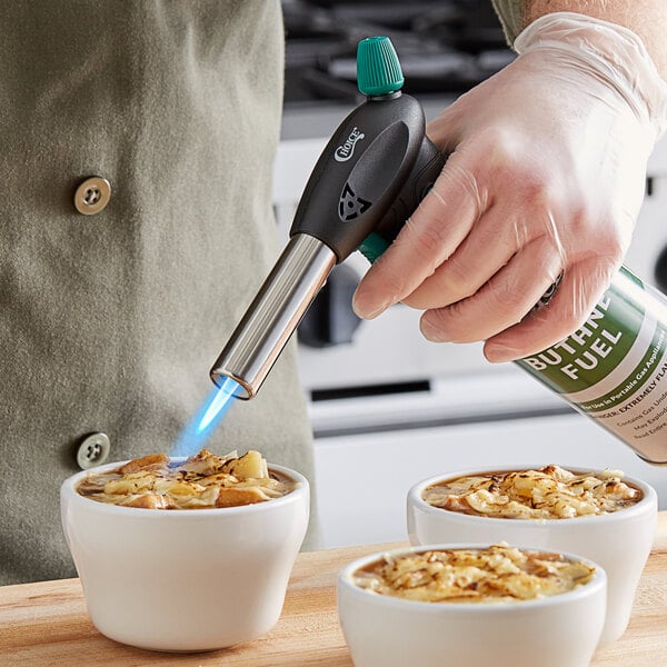 A hand holding a green Choice Butane Fuel canister over a white cup of soup.