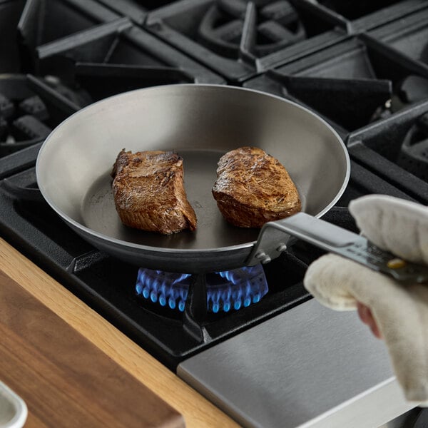 A person cooking steak in a de Buyer carbon steel fry pan on a stove.