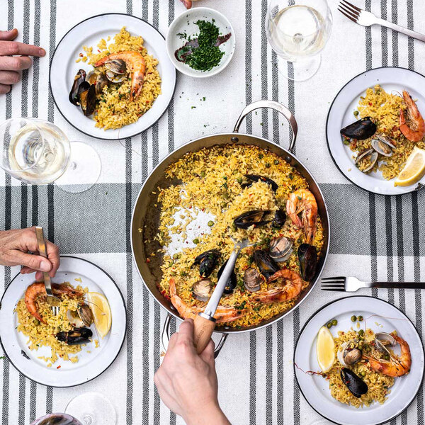 A de Buyer paella pan filled with rice, seafood, and a spoon on a table with a glass of wine.