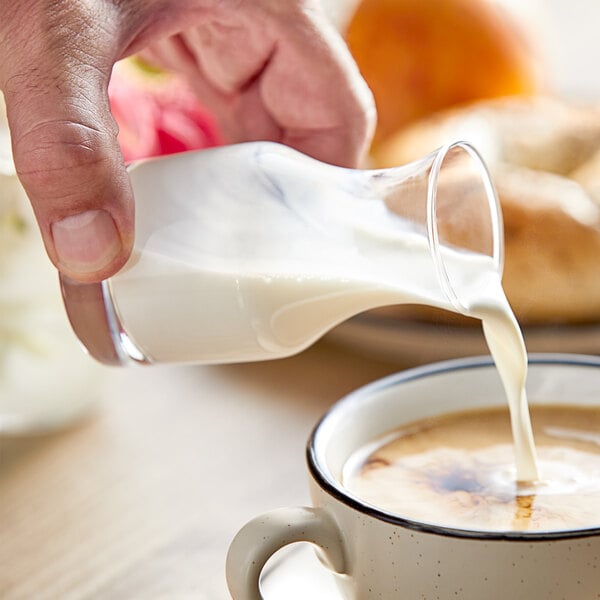 A hand pouring milk from an Acopa glass carafe into a cup of coffee.