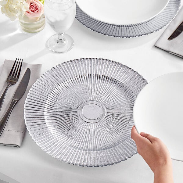 A hand reaching for an Acopa silver sunburst charger plate on a table with a knife and glass of water.