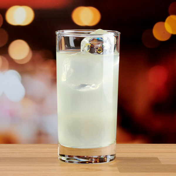 A clear 12 oz. highball glass filled with a light-colored beverage and ice, placed on a wooden surface.