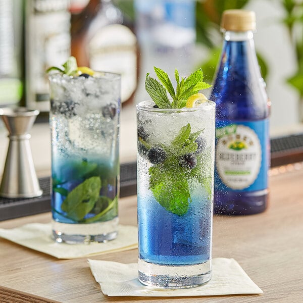 Two glasses of blueberry syrup cocktails with mint leaves on a table.