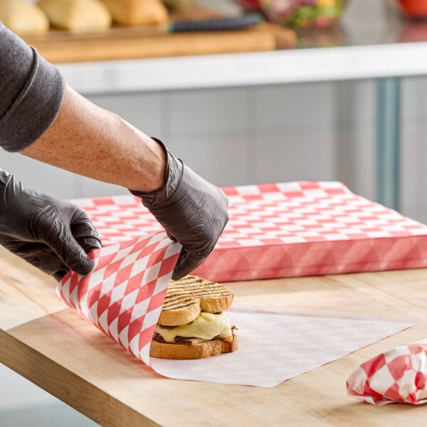 A sheet of red check deli sandwich wrap paper being used to wrap a sandwich on a wooden counter.