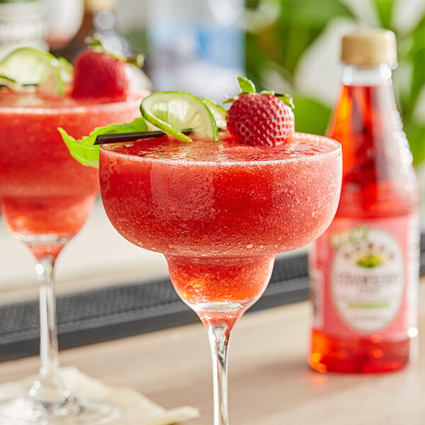 Two glasses of strawberry margaritas garnished with strawberries and limes made with Rose's Strawberry Syrup on a table in a cocktail bar.