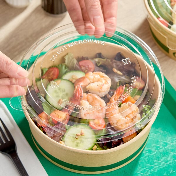 A clear round compostable PLA take-out lid being placed on a paper food container filled with salad and shrimp.