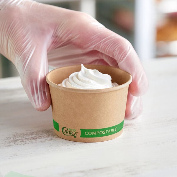 A small round kraft compostable take-out container with a swirl of whipped topping, being held by a gloved hand.