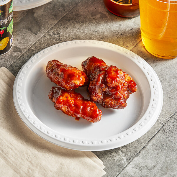 A Huhtamaki Chinet white plastic plate with chicken wings and sauce on a table with a drink.