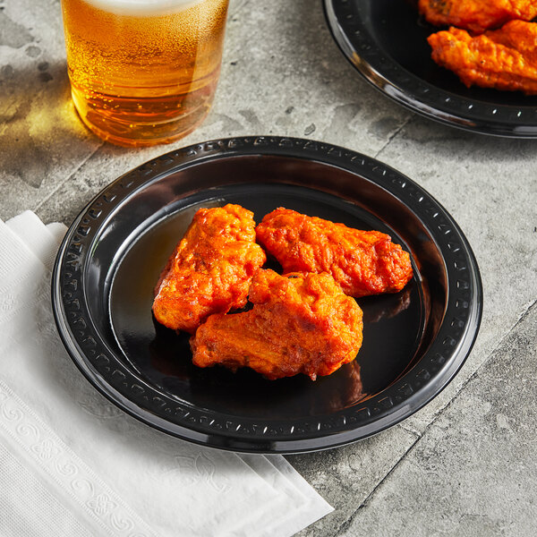 A Huhtamaki Chinet black plastic plate with chicken wings and a glass of beer.