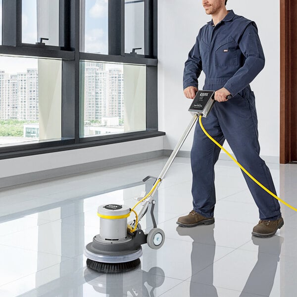 A man in blue overalls using a Lavex Pro single speed rotary floor machine to clean a floor in a professional kitchen.