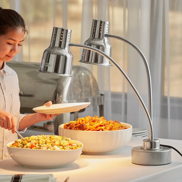 An Avantco stainless steel dual arm heat lamp over a table with a bowl of pasta and a woman holding a plate over a bowl of food.