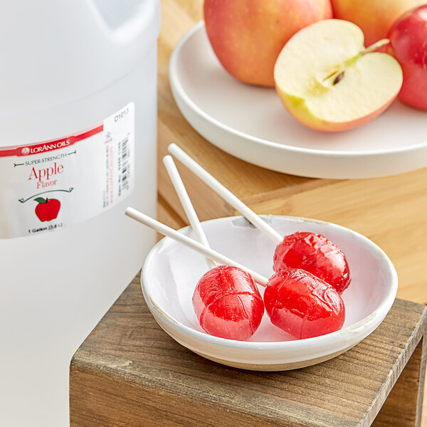 A white jug with a red label of LorAnn Oils Apple Super Strength Flavor on a table with a bowl of apples.