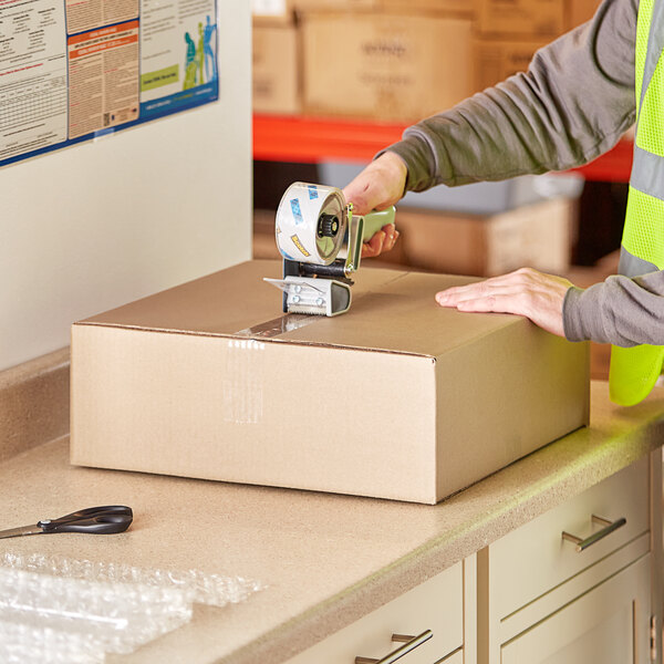 A person using a tape dispenser on a Lavex Kraft corrugated shipping box.