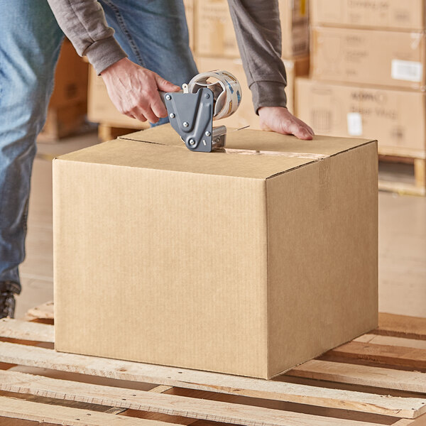 A person using a box cutter to cut a Lavex Kraft cardboard shipping box.