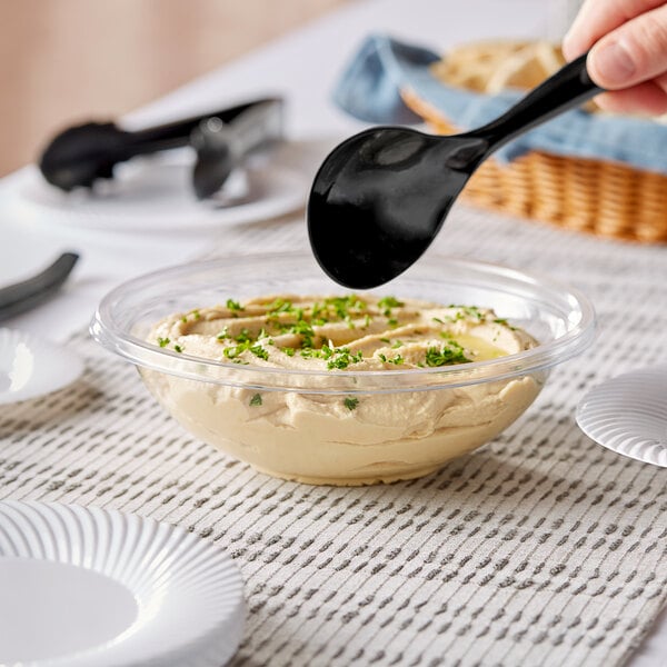 A person using a black spoon to serve hummus from a Visions clear plastic bowl.