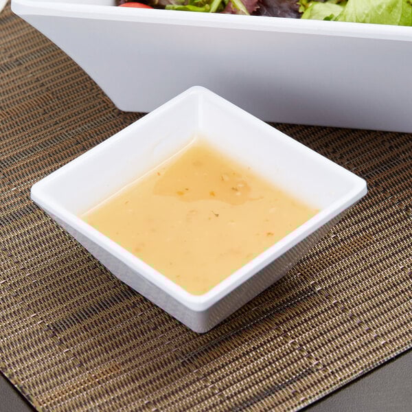 A white square GET Melamine bowl with salad next to a bowl of dressing.