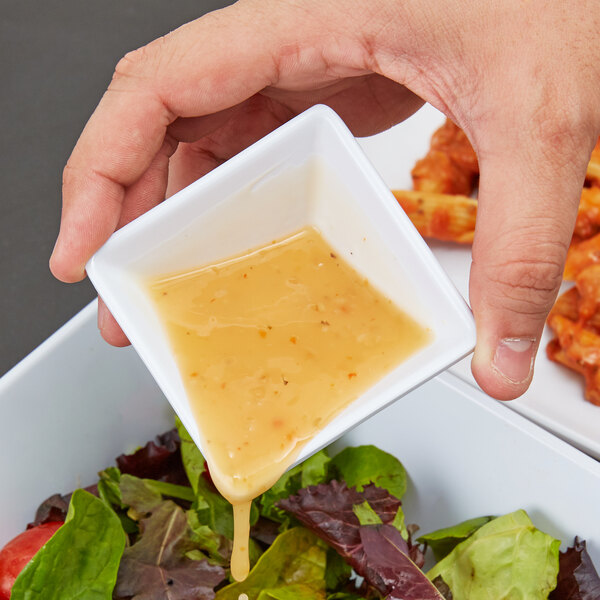 A hand holding a square white bowl with yellow sauce being poured over a salad.