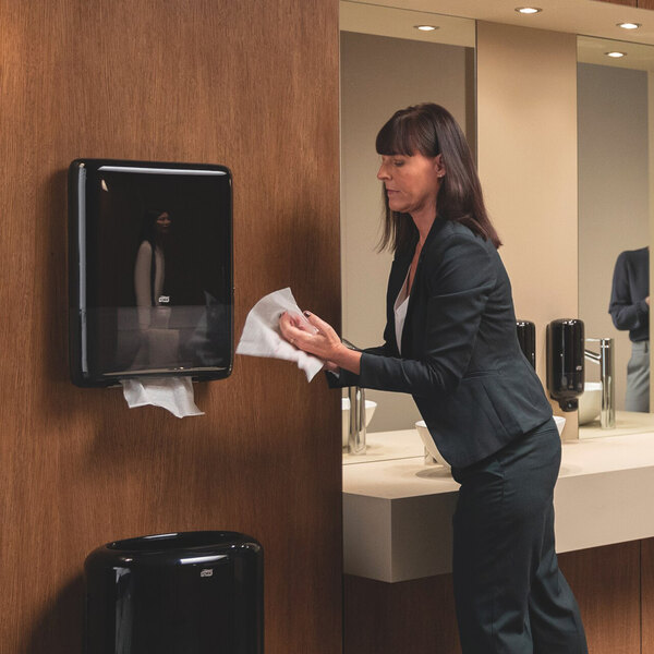 A black Tork Peakserve Mini Continuous Paper Towel Dispenser mounted on a restroom wall.