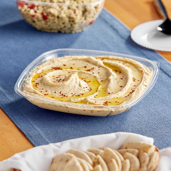 A Visions clear plastic square bowl filled with hummus and crackers on a table.