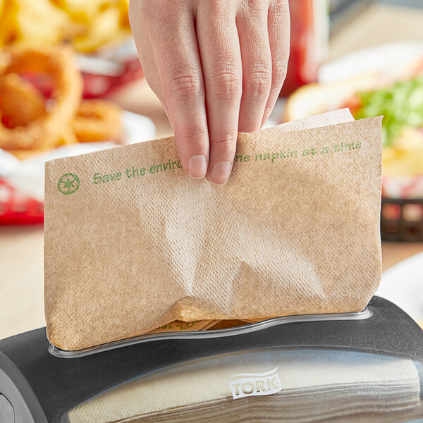 A hand holding a Tork dispenser napkin with green environmental print in a dispenser