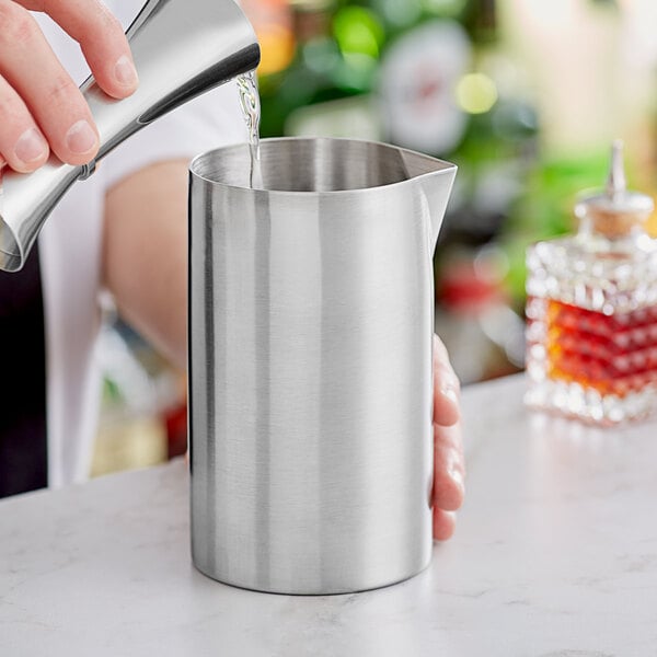 A hand pouring liquid into a silver Barfly stainless steel mixing tin.