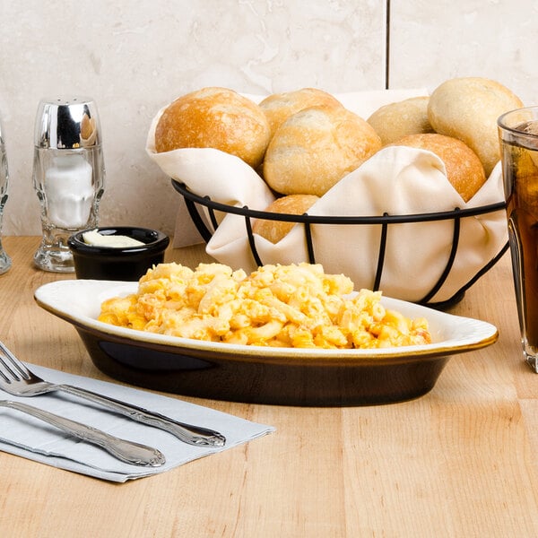 A black Tuxton oval dish with macaroni and cheese on a table with a glass of soda and a white object in it.