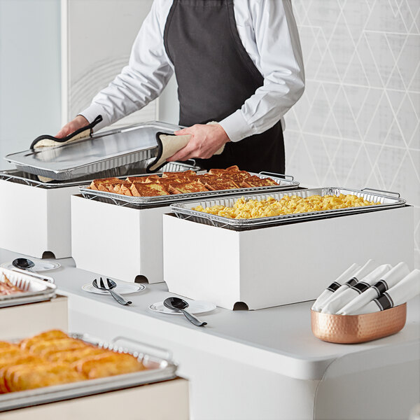 A man wearing a black apron holding a tray of food over a white tablecloth with a bowl and utensils on it.