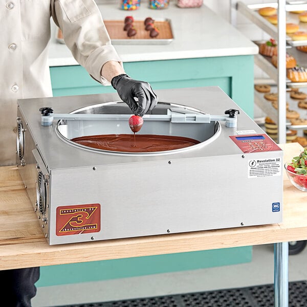 A person in a white coat and black gloves using a ChocoVision Revolation 3Z chocolate tempering machine to pour chocolate over a dessert.
